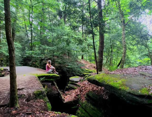 Lady sitting on a rock ledge with green moss on it, overlooking a trail down below.