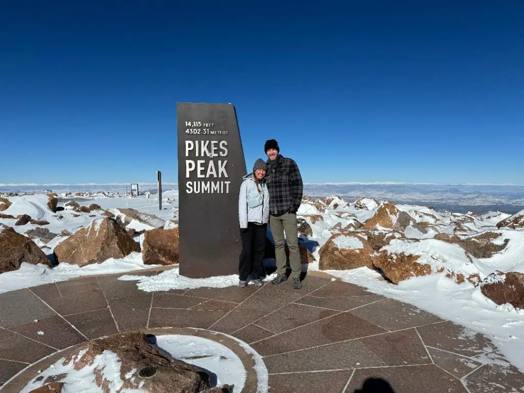 couple standing in front of Pikes Peak Summit Sign on a snowy covered mountain top at Pikes Peak