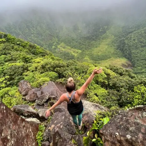 girl standing on a mountain ledge over green rainforest and a volcano during while hiking in st kitts