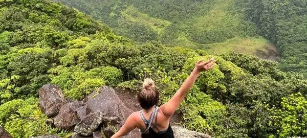 girl standing on a mountain ledge over green rainforest and a volcano during while hiking in st kitts