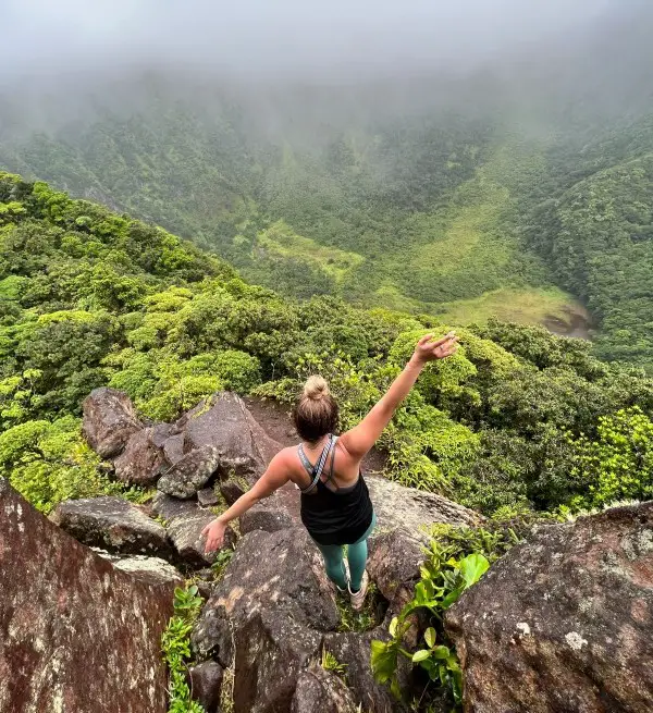 girl standing on a mountain ledge over green rainforest and a volcano during while hiking in st kitts