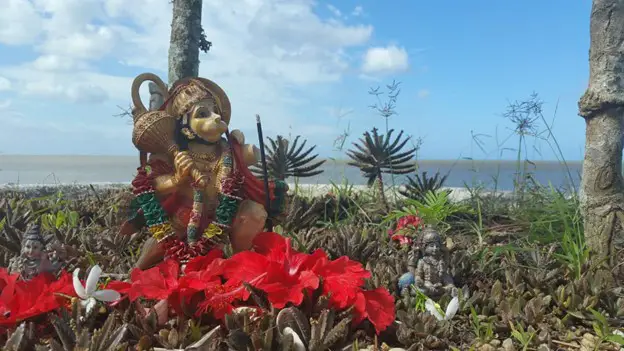 behind some red flowers is a golden statue Deity figure at Temple in the Sea in Trinidad