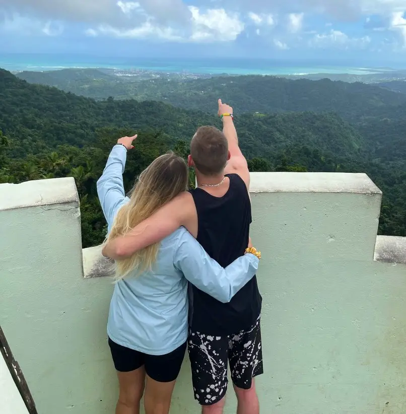 couple with arms around each other point in the distance to the blue water over the trees on the horizon