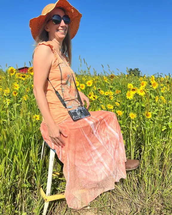 lady sitting on a stool in a field of yellow sunflowers with an orange hat and a cool camera phone case