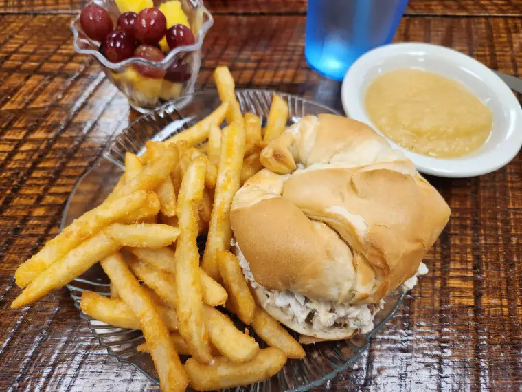 a plate with a crescent roll bun with salad on it and fries, bowl of fruit, and bowl of applesauce on a wooden table