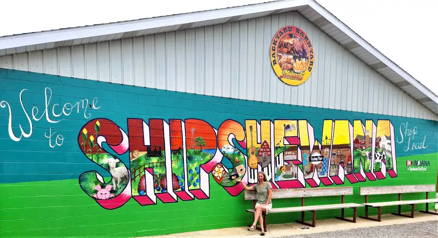 lady sitting on a bench in front of a bright colored SHIPSHEWANA mural on a white building