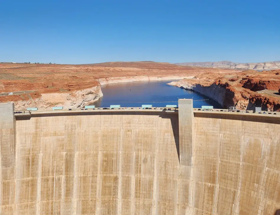 view of the top of a large dam with a reservoir in the backof it and desert landscape stretched out beyond that.