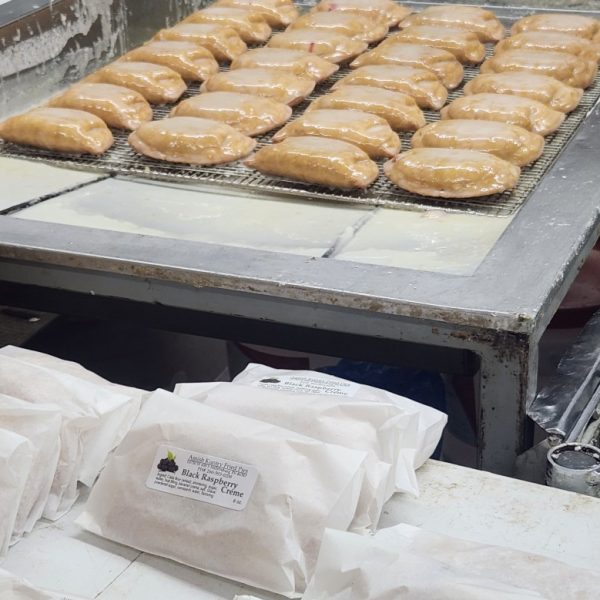 wrapped pies for sale in front of a large rack of cooling handmade pies with glaze on top