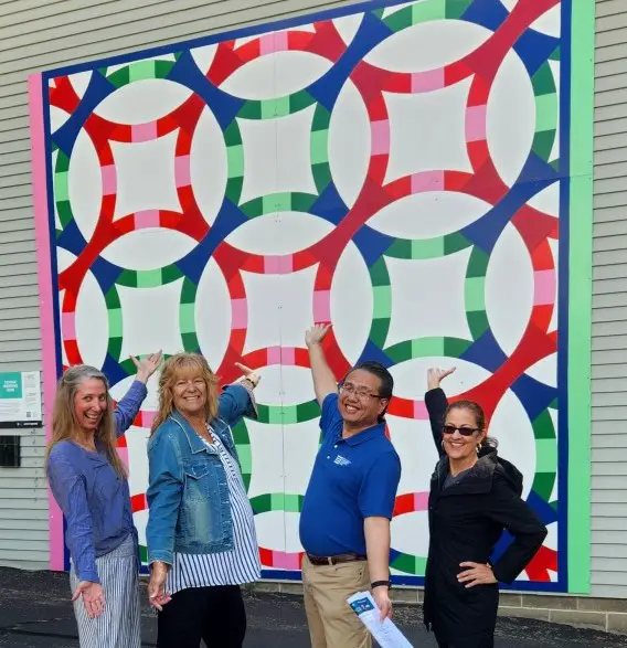 4 people with outstretched arms standing in front of a colorful quilt mural on a wall.