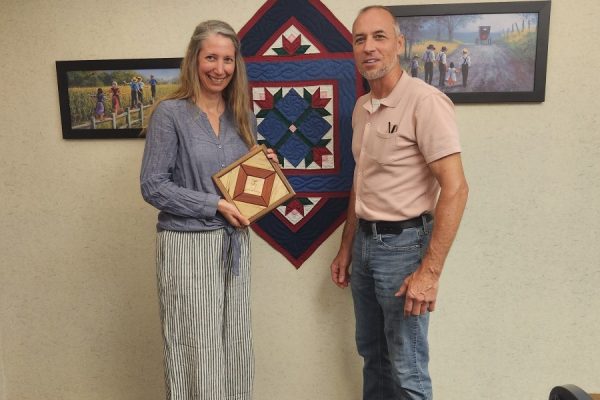 lady holding a wooden quilt square craft with man standing in front of a quilt
