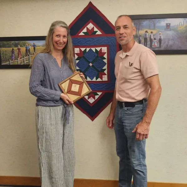 lady holding a wooden quilt square craft with man standing in front of a quilt