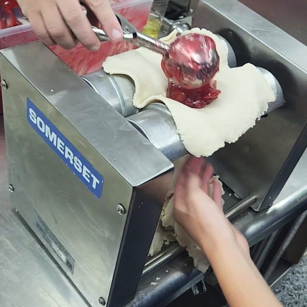 scoop of fruit being placed into a flat dough inserted into a machine to make an Amish Pie