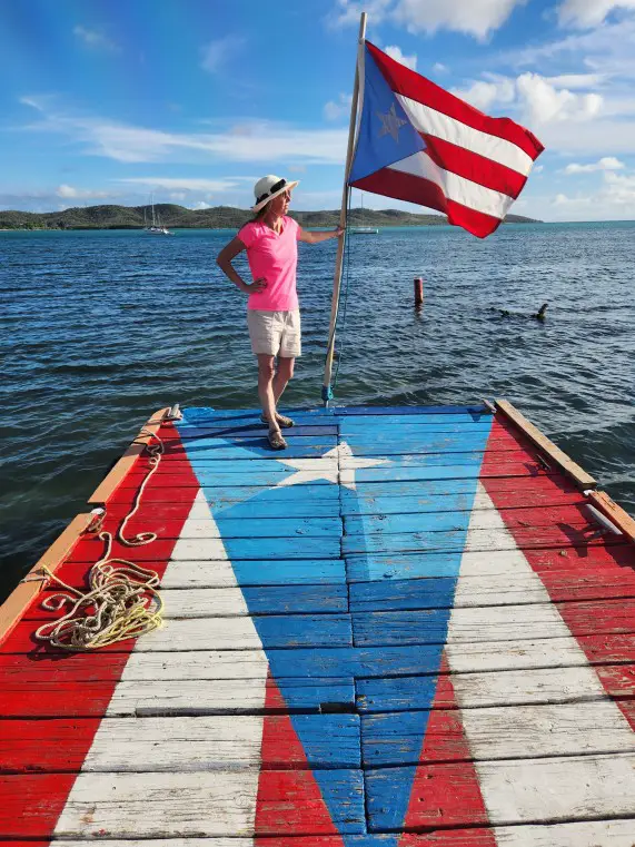 lady in bright pink shirt holding Puerto Rico flag and standing on a dock painted to resemble the Puerto Rico flag.