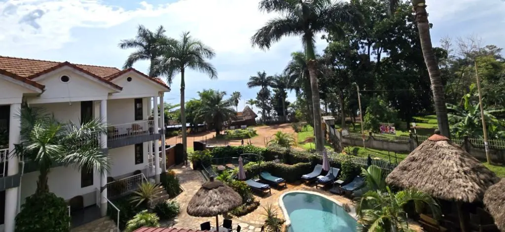 view from above of a blue pool with a white hotel in the background and palm trees and lake Victoria in the distance