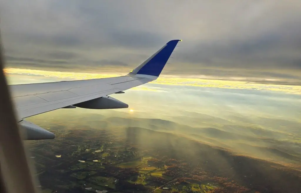 wing of airplane in the clouds with sun peaking out in the background