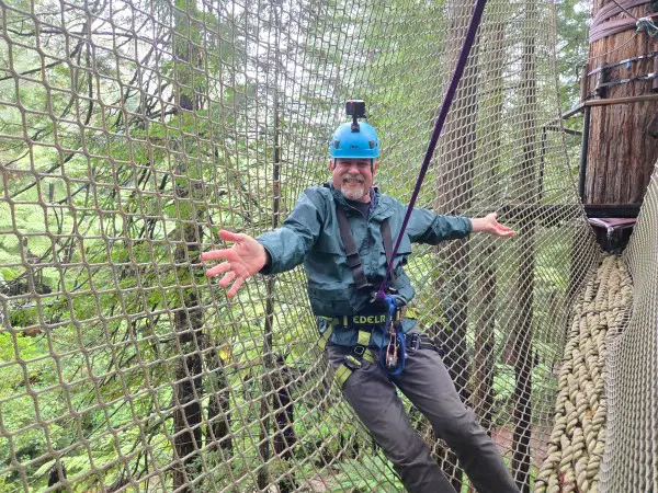 man leaning on a rope bridge above a forest of redwood trees in new zealand