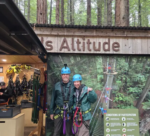 couple standing together in front of Altitude sign with zip line gear, carabiners, and harnesses