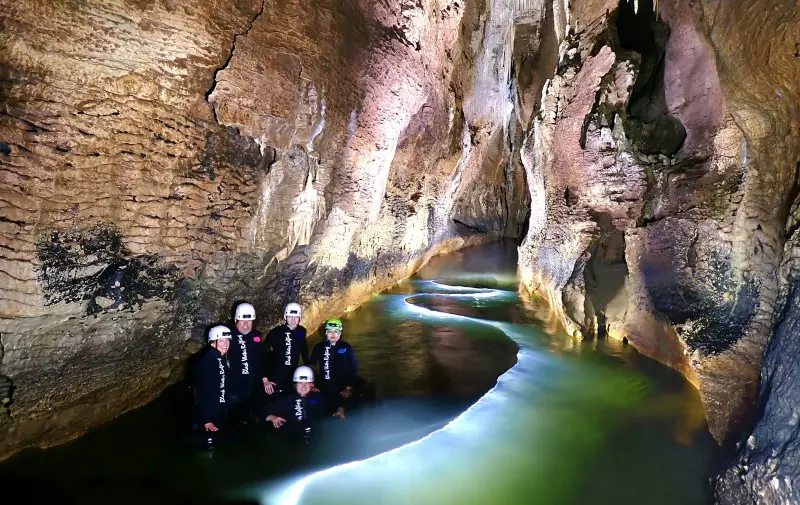 group of people in a dimly lit cave, standing in water with helmets and a blue glowing light through the water near Rotorua New Zealand