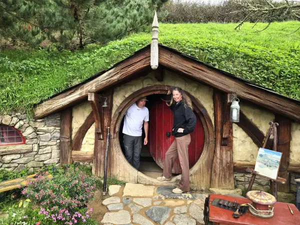 man and woman standing in the doorway of a small circular door of a hobbit house in Hobbiton New Zealand