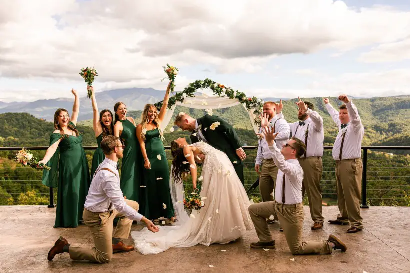wedding party celebrating and throwing flowers and their hands up in the air while groom dips the bride in a smoky mountain scenery backdrop and white cloudy skies