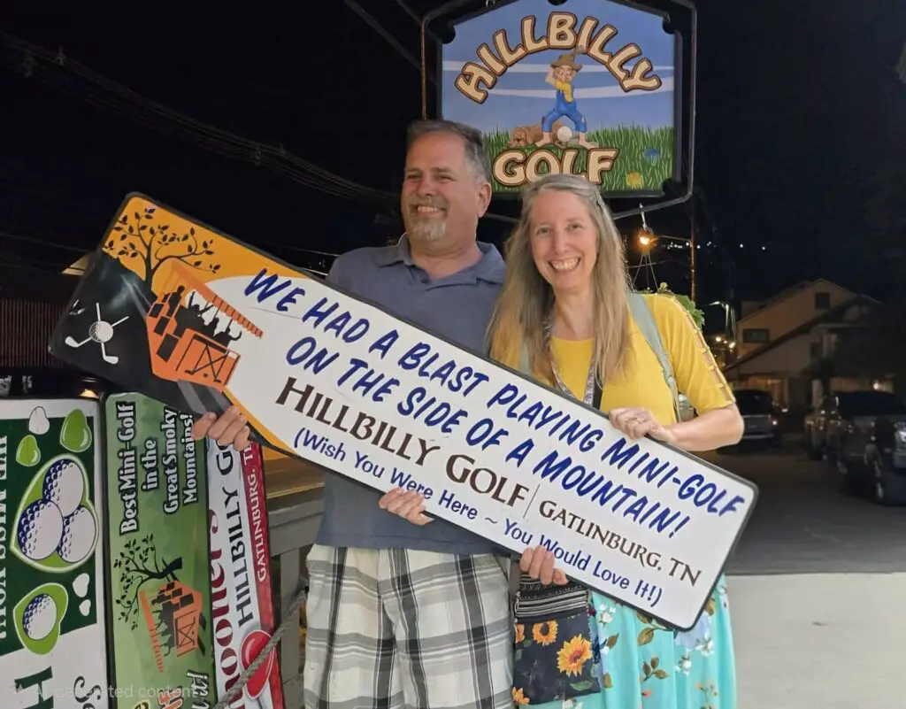 man and woman holding a sign that says We had a blast playing mini-golf on the side of a mountain HILLBILLY GOLF