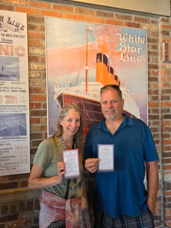 couple holding up a personal story of a passenget on the Titanic in front of a poster of the Titanic Ship